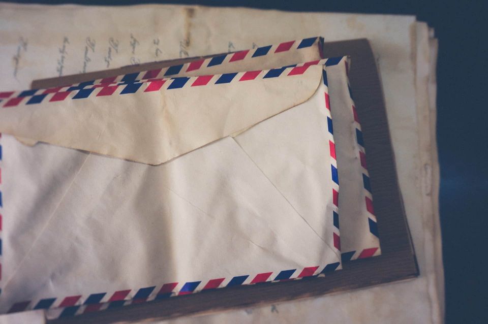 Imagem com 3 envelopes de cartas sobrepostos, eles tem listras azuis e vermelhas e abaixo deles estão folhas envelhecidas.