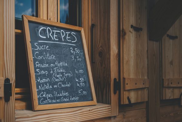 Imagem com tons escuros. Lousa com preços de crepes, está em inglês. Ao fundo uma janela de madeira.